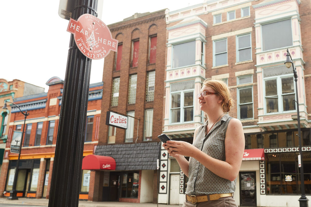 woman using phone looking at Hear, Here sign